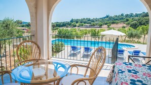 En balkon med et glasbord og stole, med udsigt over en swimmingpool og frodig vegetation.