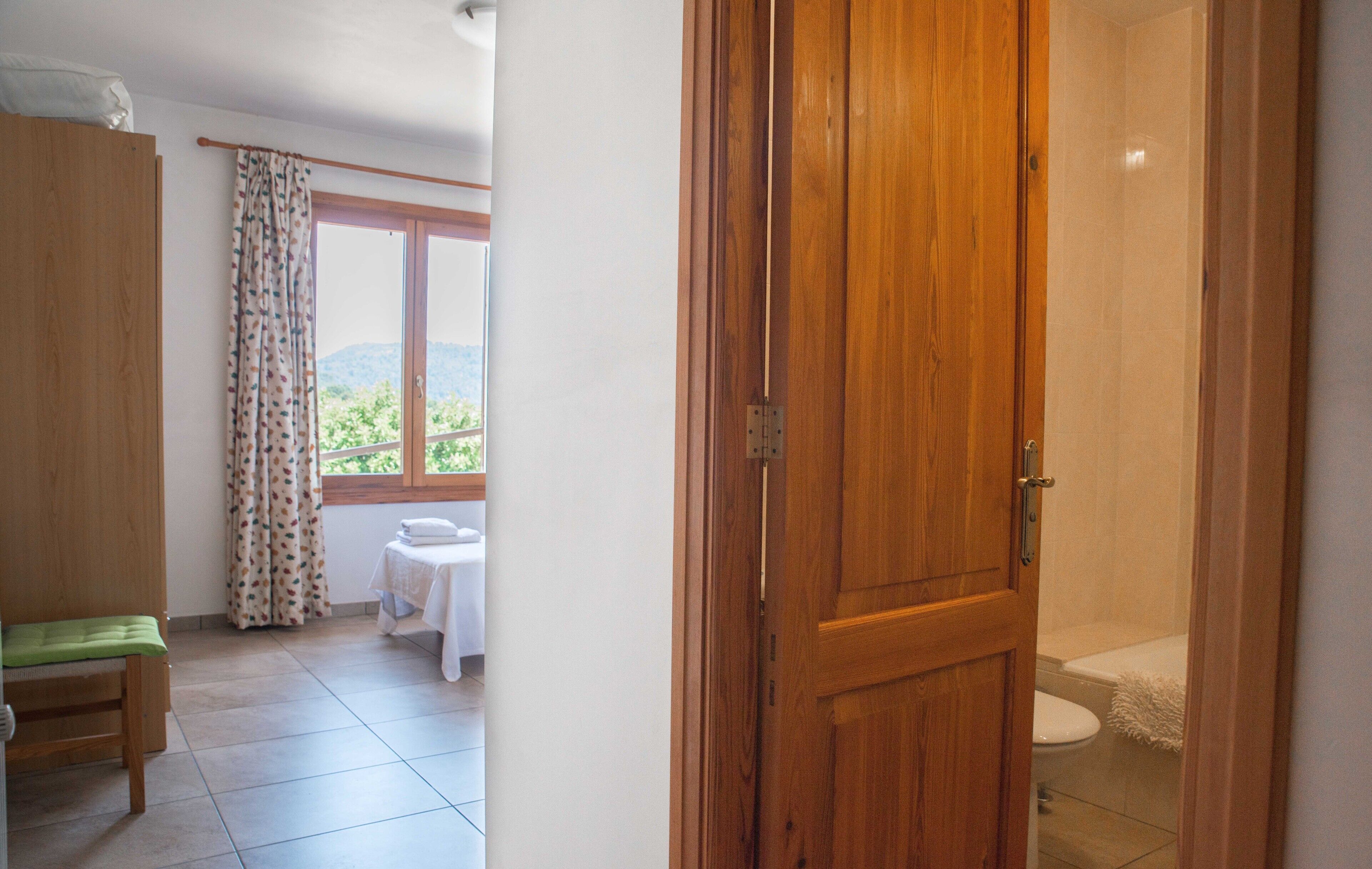 Ein Zimmer mit Holztür, einer Bank, einem kleinen Tisch mit weißem Tuch und Blick ins Grüne durch ein Fenster.
