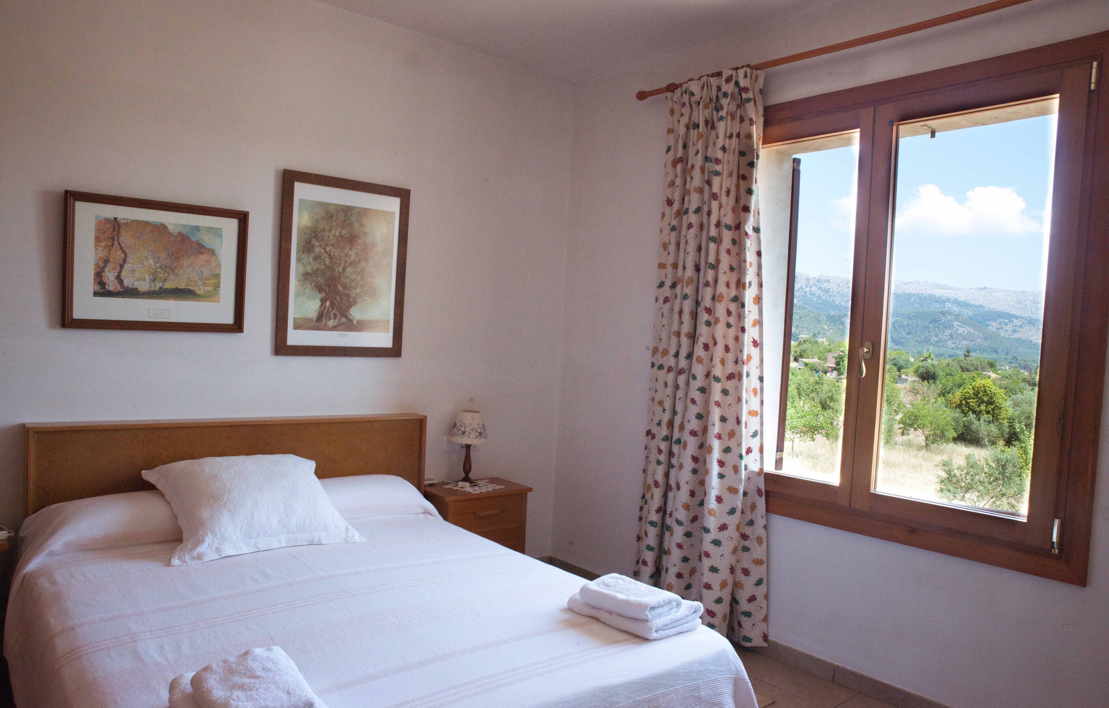 A bedroom with a bed, two framed artworks, a nightstand, a lamp, and a window with a view of the landscape.