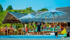 A poolside area with lounge chairs and umbrellas, a person serving drinks, and a thatched-roof building in the background. - Emeraude Lodge (Nosy Be)