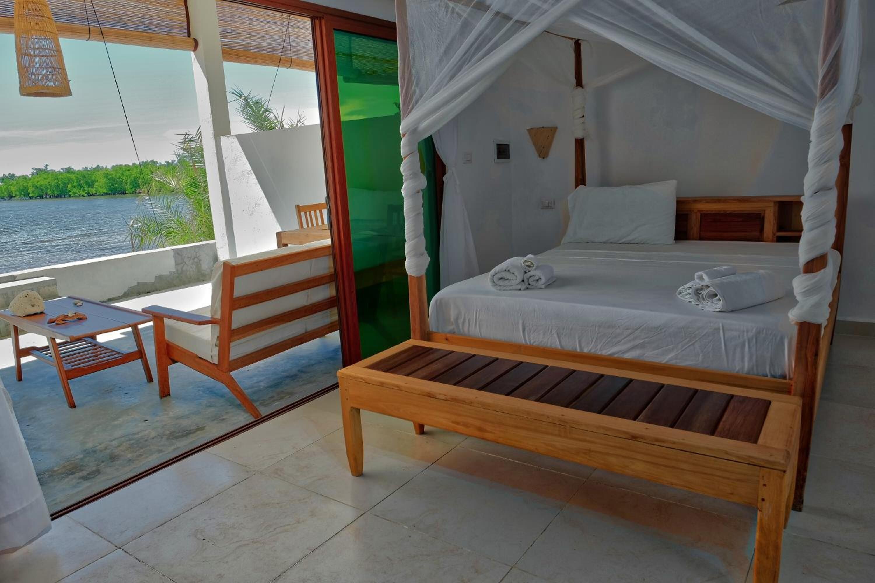 Een slaapkamer met een houten bed, wit beddengoed, een houten bankje en uitzicht op een waterpartij.