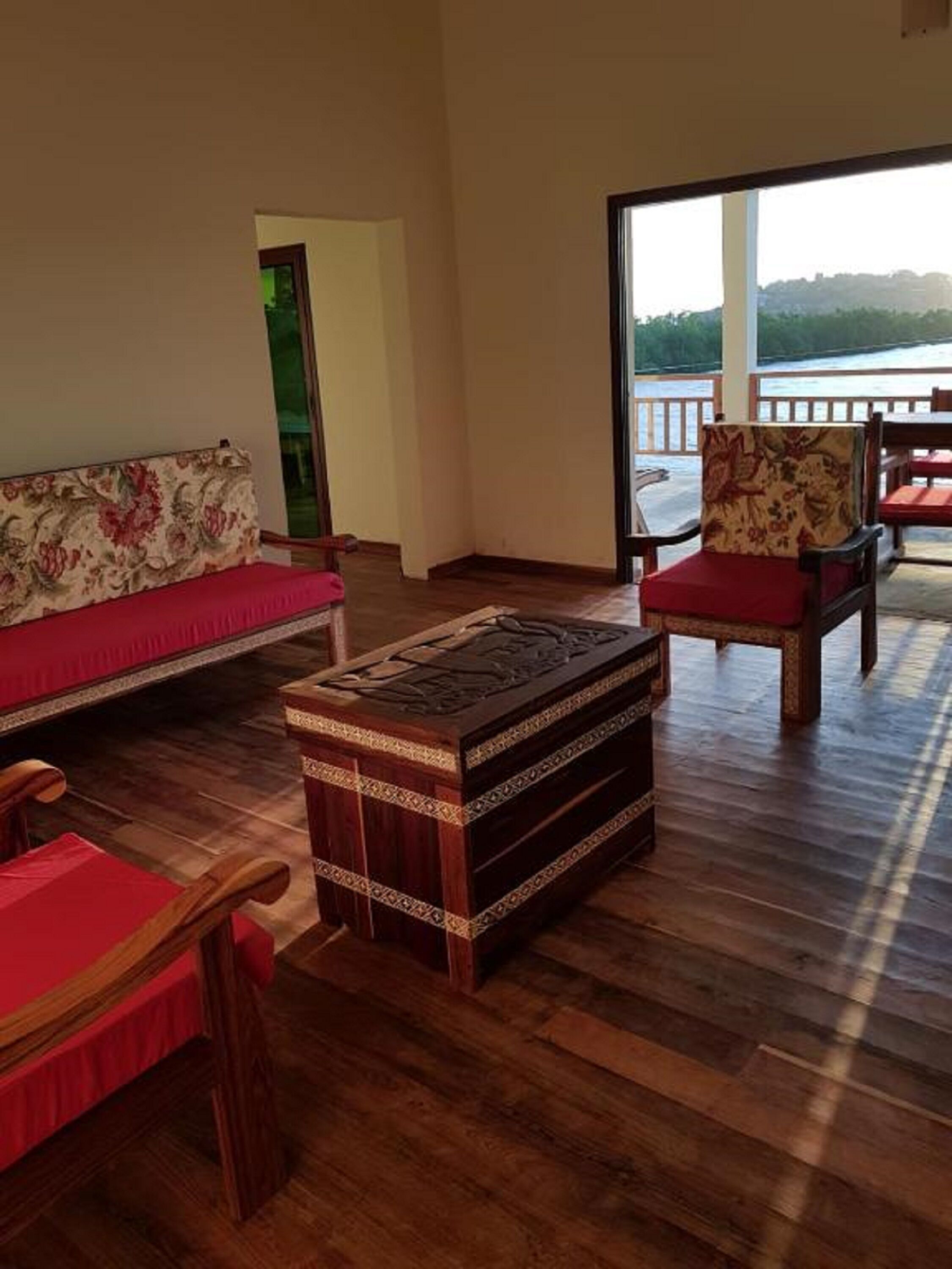 A room with a wooden floor, a sofa with a floral pattern, a wooden chest of drawers, and a chair with a red cushion.