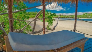 A wooden lounge chair with a cushion under a thatched roof, overlooking a beach. - Emeraude Lodge (Nosy Be)