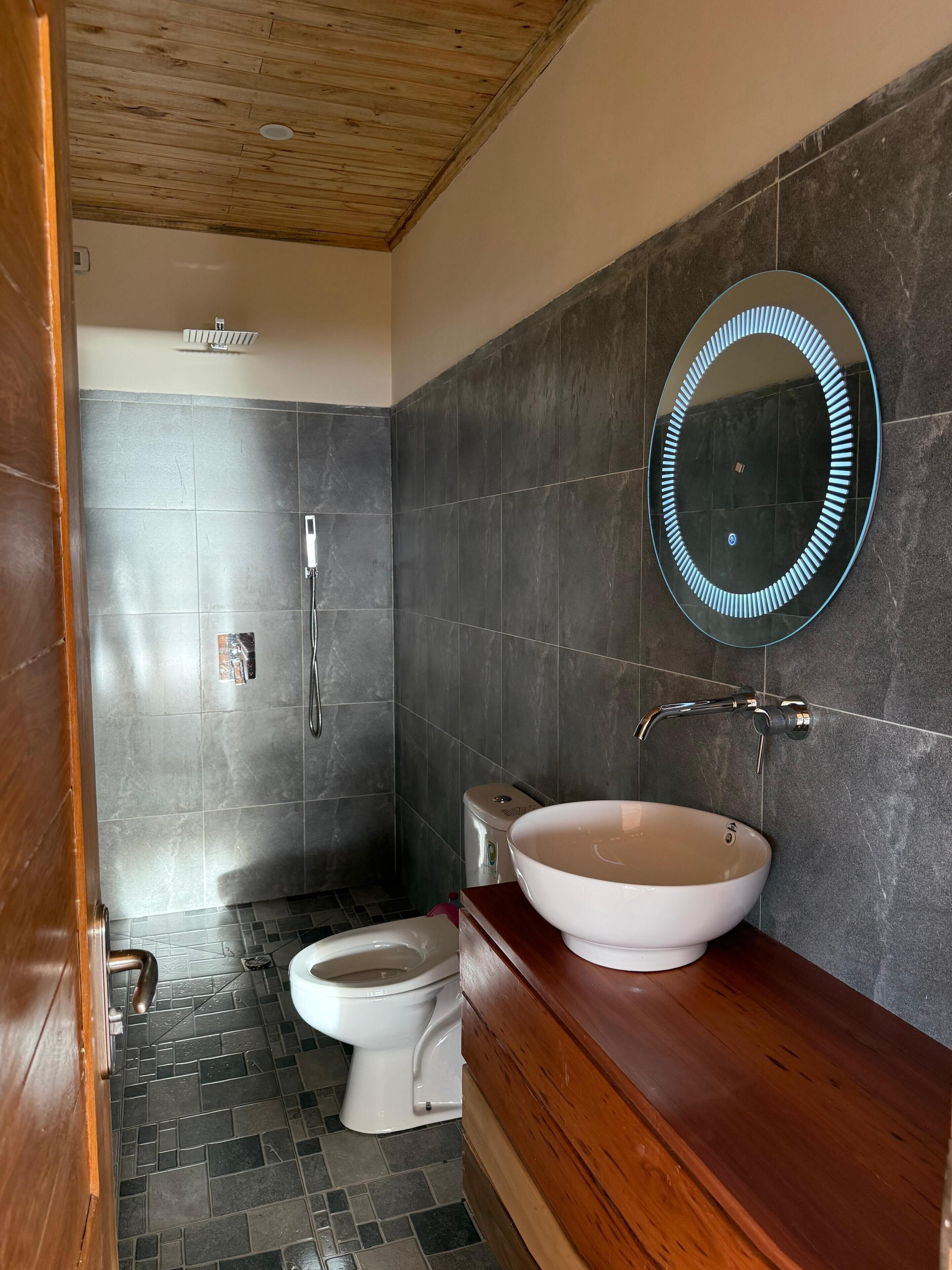 A bathroom with a wooden vanity, a round mirror, a white sink, and a shower.