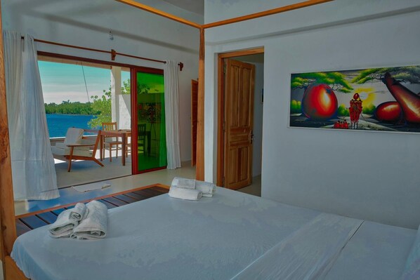A hotel room with a bed, a painting, a view of the sea, and a balcony with a table and chairs. - Emeraude Lodge (Nosy Be)