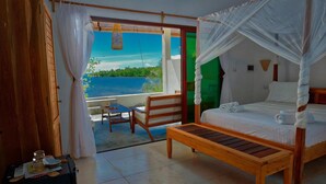 A room with a bed, a wooden bench, a table, and a view of a lake. - Emeraude Lodge (Nosy Be)