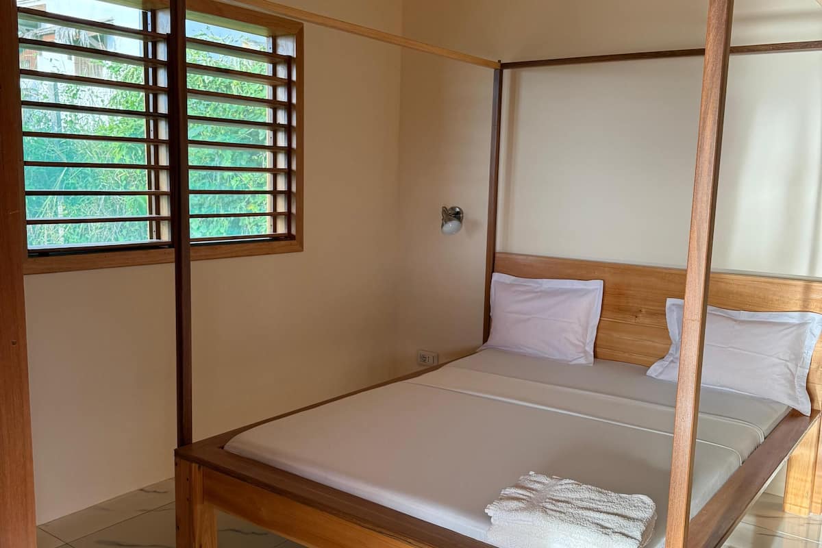 A four-poster bed with a wooden frame and white bedding in a room with a tiled floor and a window with blinds.