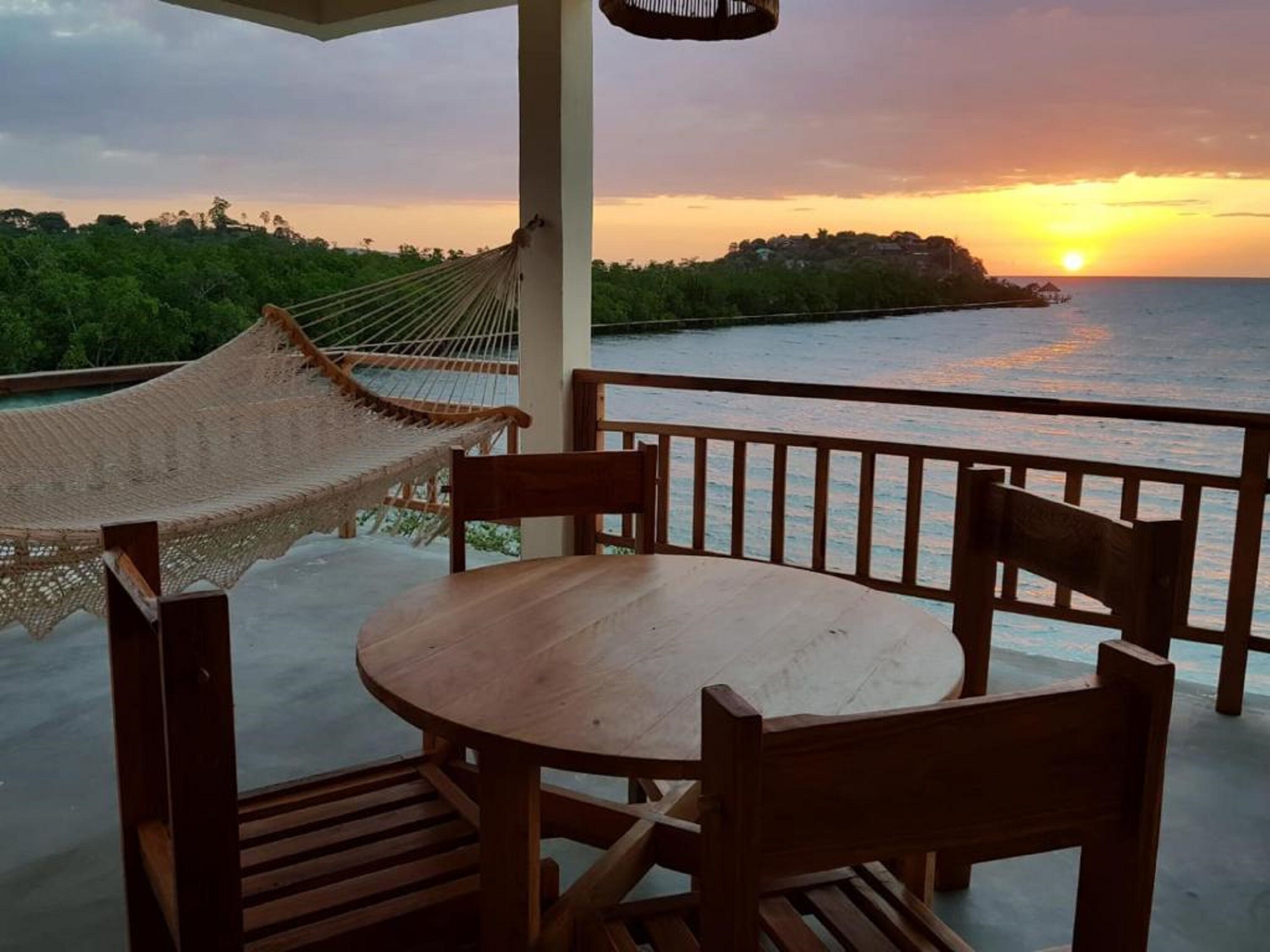 A wooden table and chairs on a balcony with a view of the sunset over the ocean.