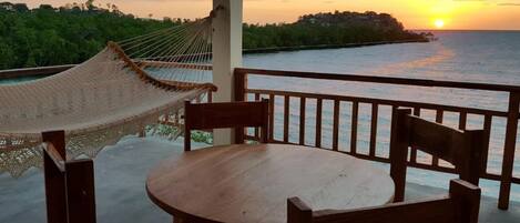 A wooden table and chairs on a balcony with a view of the sunset over the ocean.