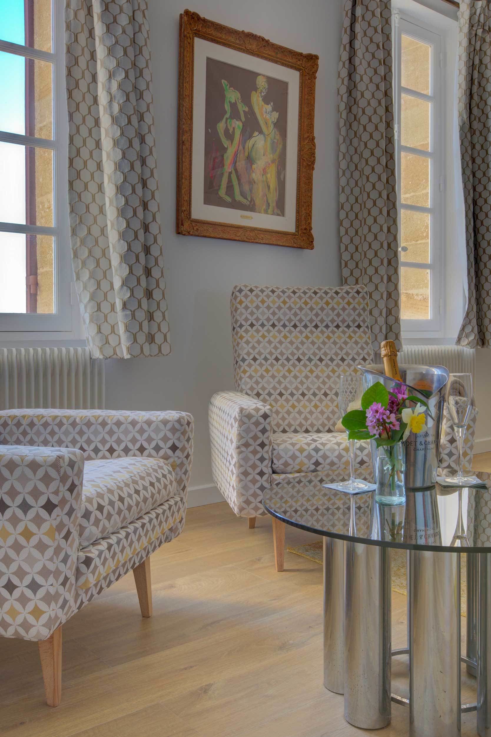 Une pièce avec des canapés à motifs, une table en verre sur laquelle se trouvent une bouteille de champagne et des fleurs, et une œuvre d’art encadrée accrochée au mur.