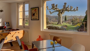 A room with a foosball table, a table with a vase of flowers, a window with a view of a tree and landscape, and a poster on the wall. - Chateau du Palanquey & SPA (Sainte-Colombe)