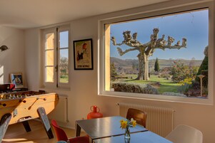 Una habitación con una mesa de fútbol de mesa, una mesa con un jarrón de flores, una ventana con vistas a un árbol y un paisaje, y un cartel en la pared.