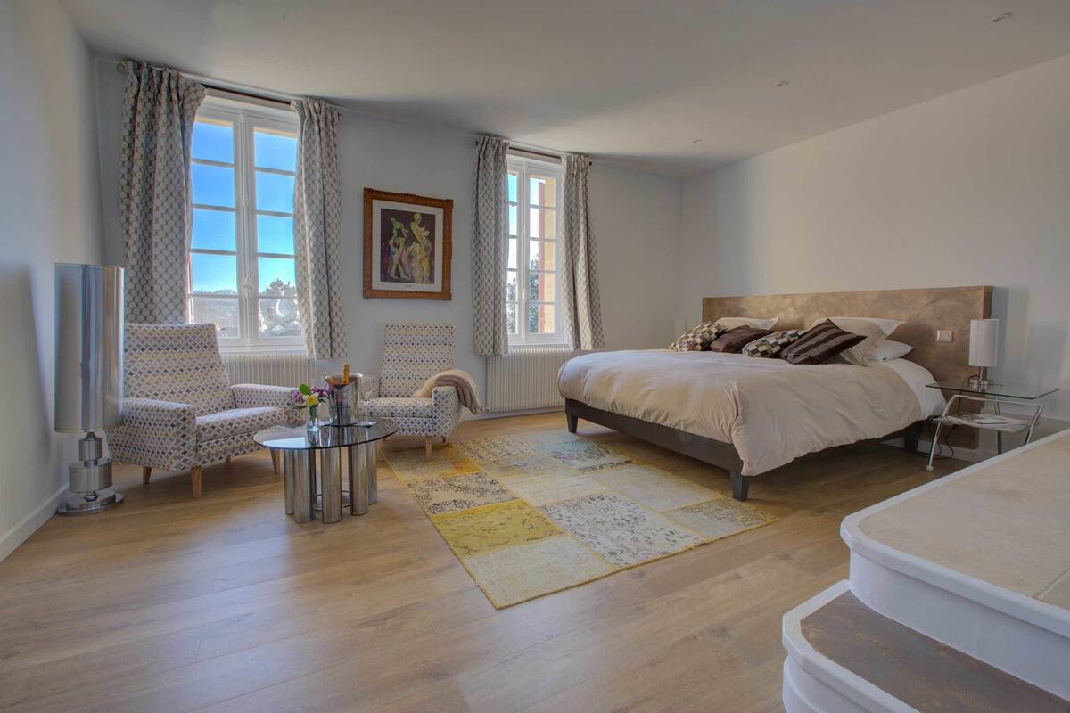 A bedroom with a large bed, a sitting area with chairs, a small table, and a window with curtains.