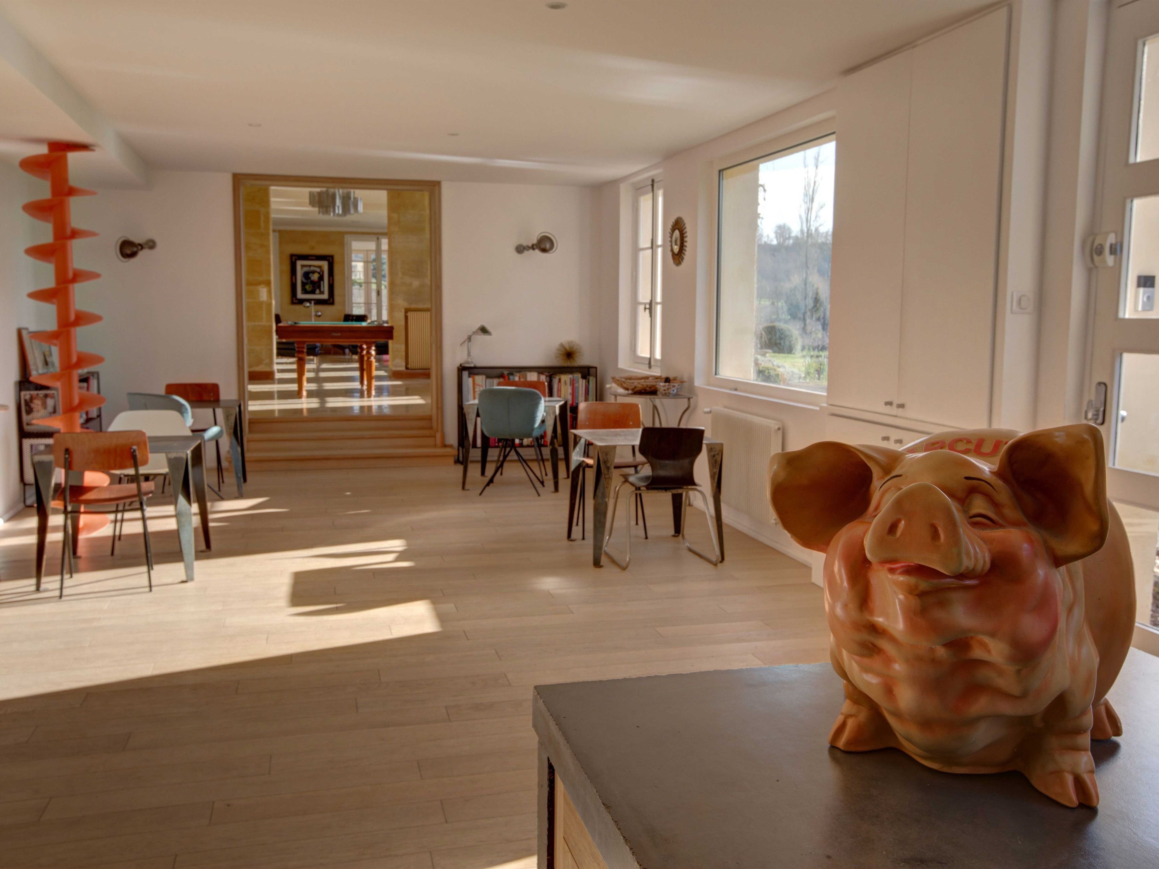 A spacious living room with a large pig sculpture, wooden flooring, and a view of the outdoors through large windows.