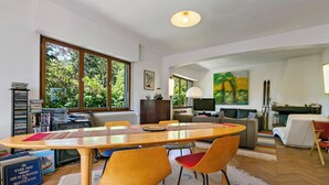 A spacious living room with a large wooden dining table, comfortable seating, a fireplace, and large windows offering a view of greenery.