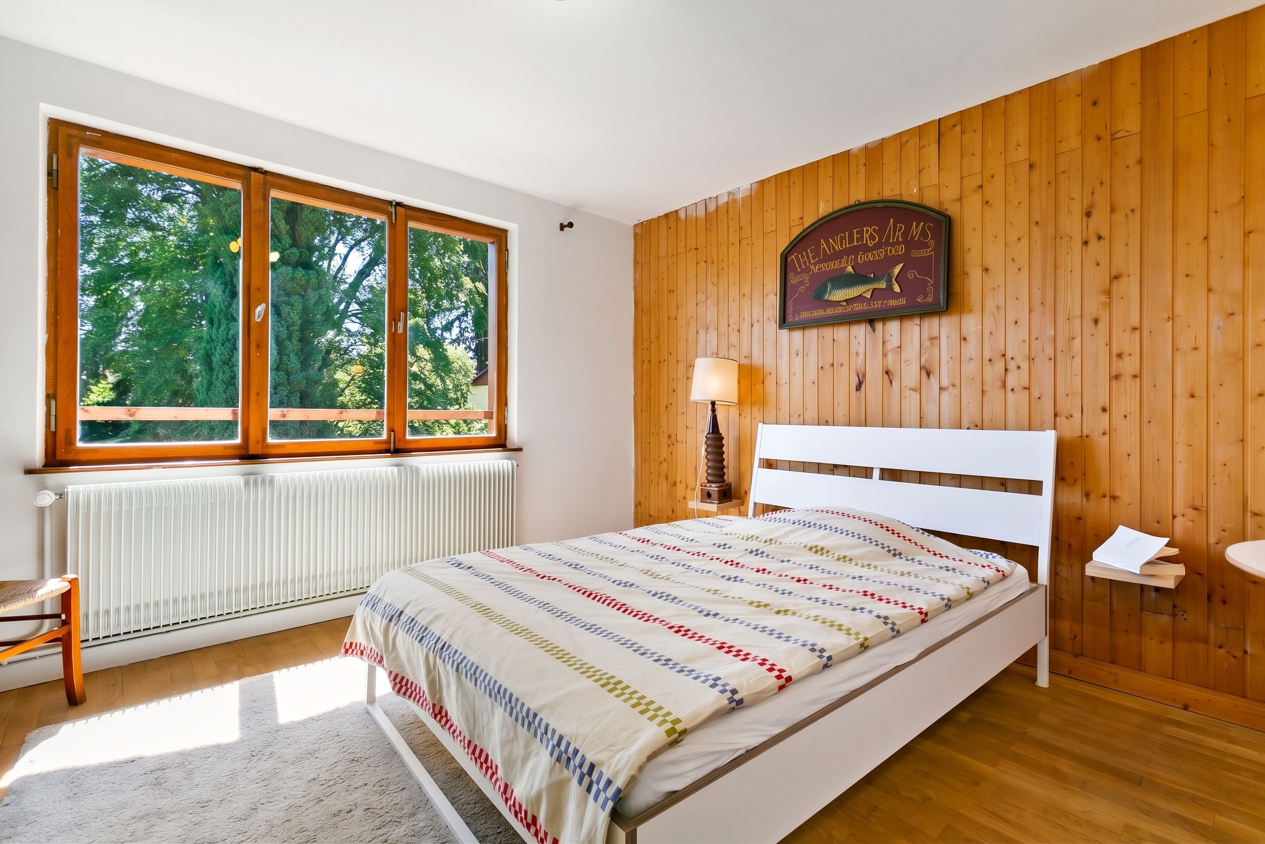 Une chambre avec une grande fenêtre, un lit recouvert d’une couvre-lit à motifs, un panneau mural en bois et un panneau indicateur placé au-dessus du lit.