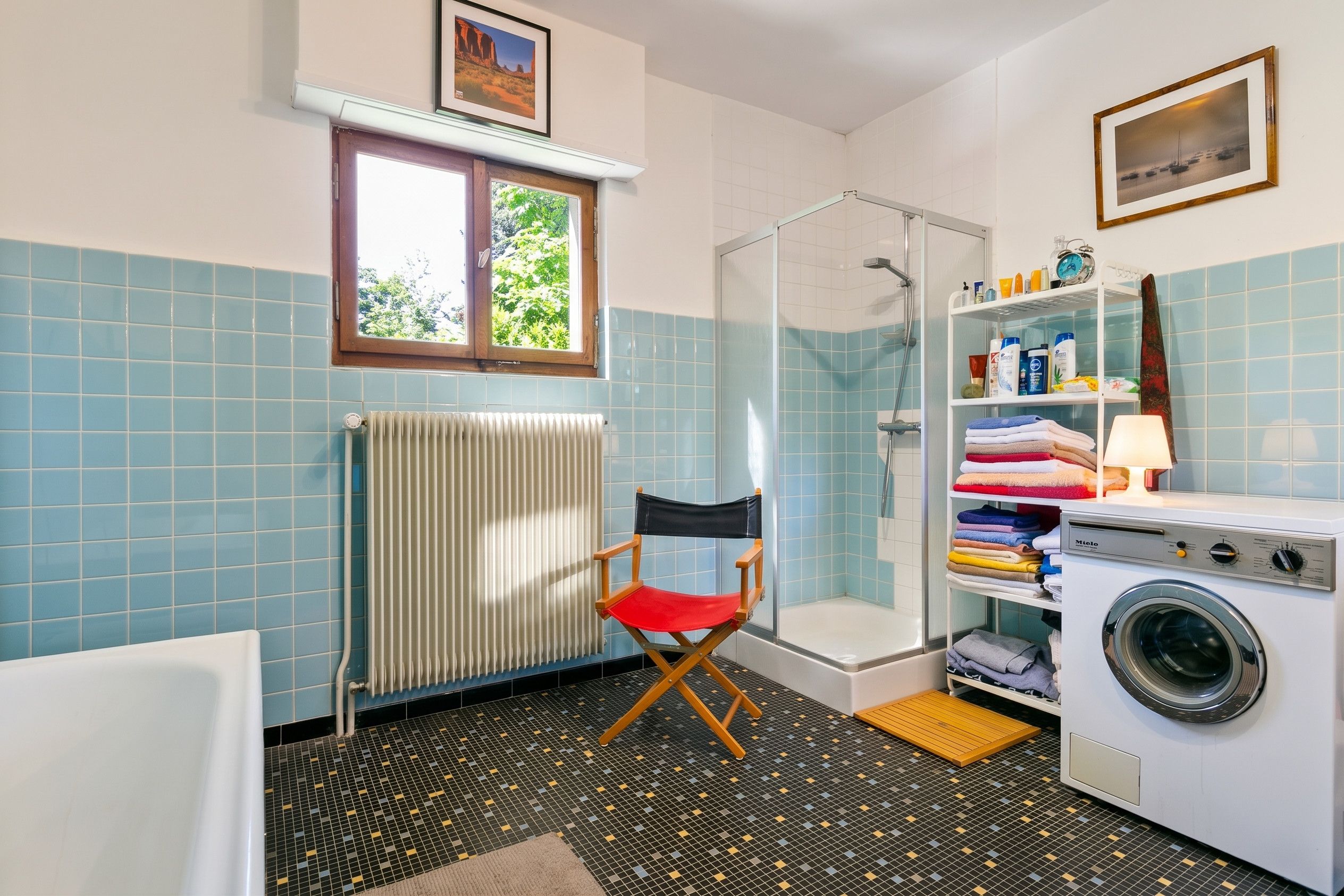 Une salle de bain équipée d’une douche, d’un radiateur, d’une fenêtre, d’une chaise, d’une étagère avec des articles de toilette et d’un lave-linge.