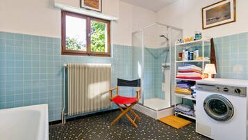 Une salle de bain équipée d’une douche, d’un radiateur, d’une fenêtre, d’une chaise, d’une étagère avec des articles de toilette et d’un lave-linge.