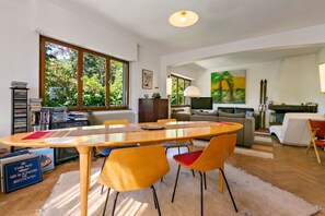 A modern living room with a dining area, large windows, and a view of greenery outside.