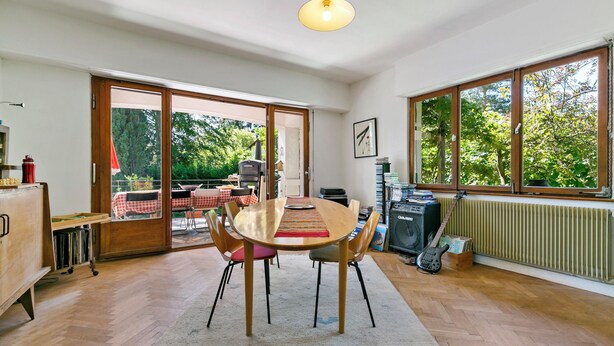 A dining area with a wooden table, chairs, and a view of the outdoors through large windows.
