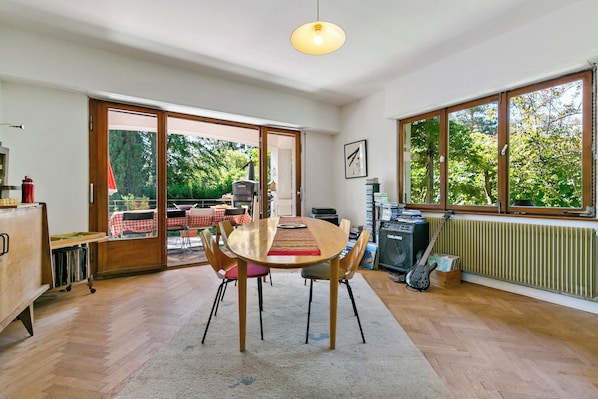 Een eetgedeelte met een houten tafel, stoelen en uitzicht op de omgeving door grote ramen.