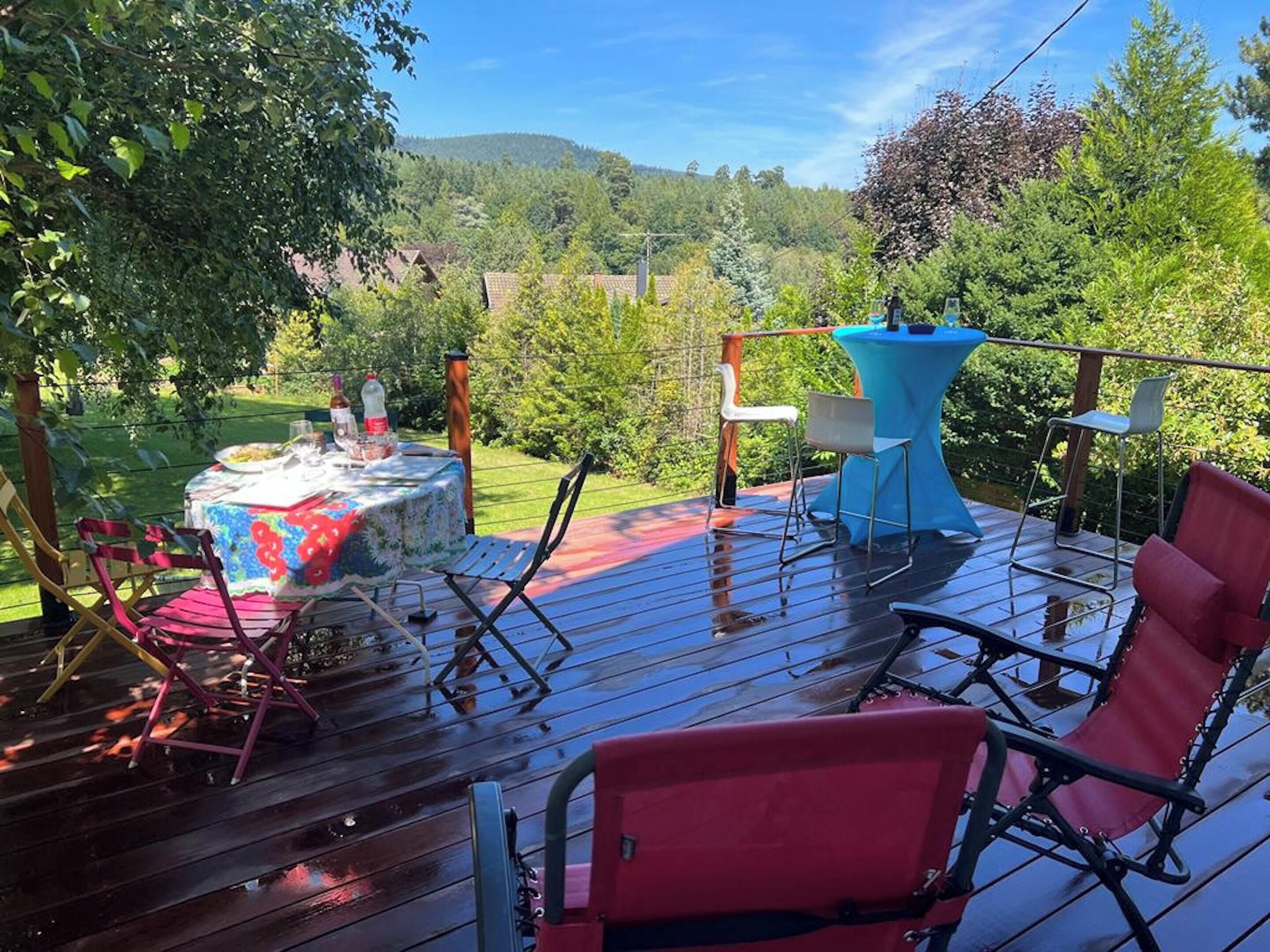 Eine Holzdiele mit Esstisch und Stühlen, mit Blick auf einen üppigen Garten und sanfte Hügel in der Ferne.