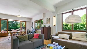 A modern living room with a grey sofa, a smaller sofa, a coffee table with books, and large windows offering a view of greenery outside.