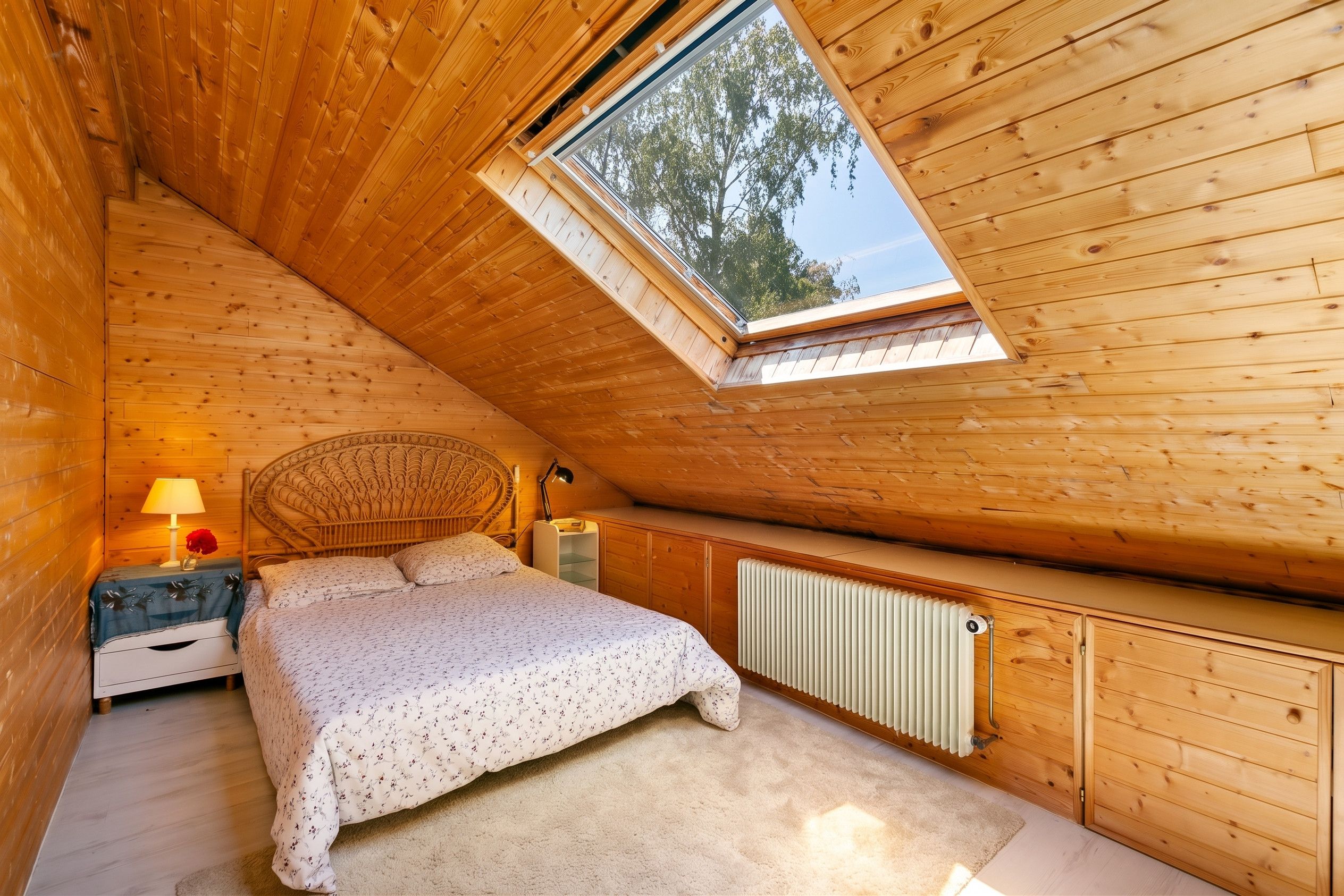 A cozy attic bedroom with a sloped ceiling, a skylight, a bed with floral bedding, a bedside lamp, a nightstand, and a radiator.