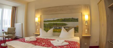 A hotel room with a large bed, two bedside lamps, a painting of a lake, a chair, and a small table.