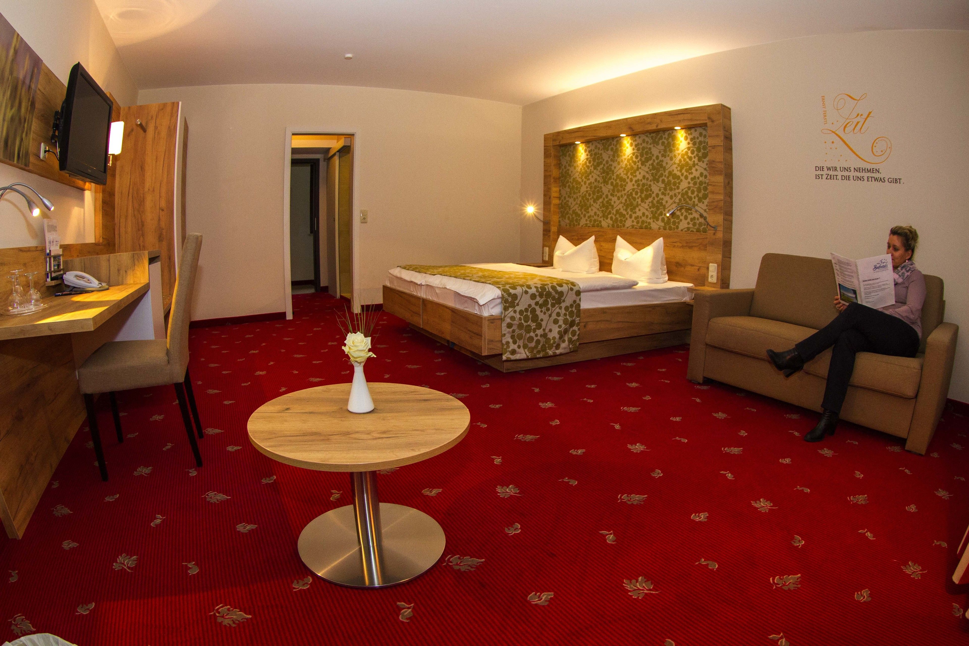 Ein Hotelzimmer mit Bett, Schreibtisch mit Telefon, Stuhl, rundem Tisch mit Vase sowie Wand mit Dekoration und Text.