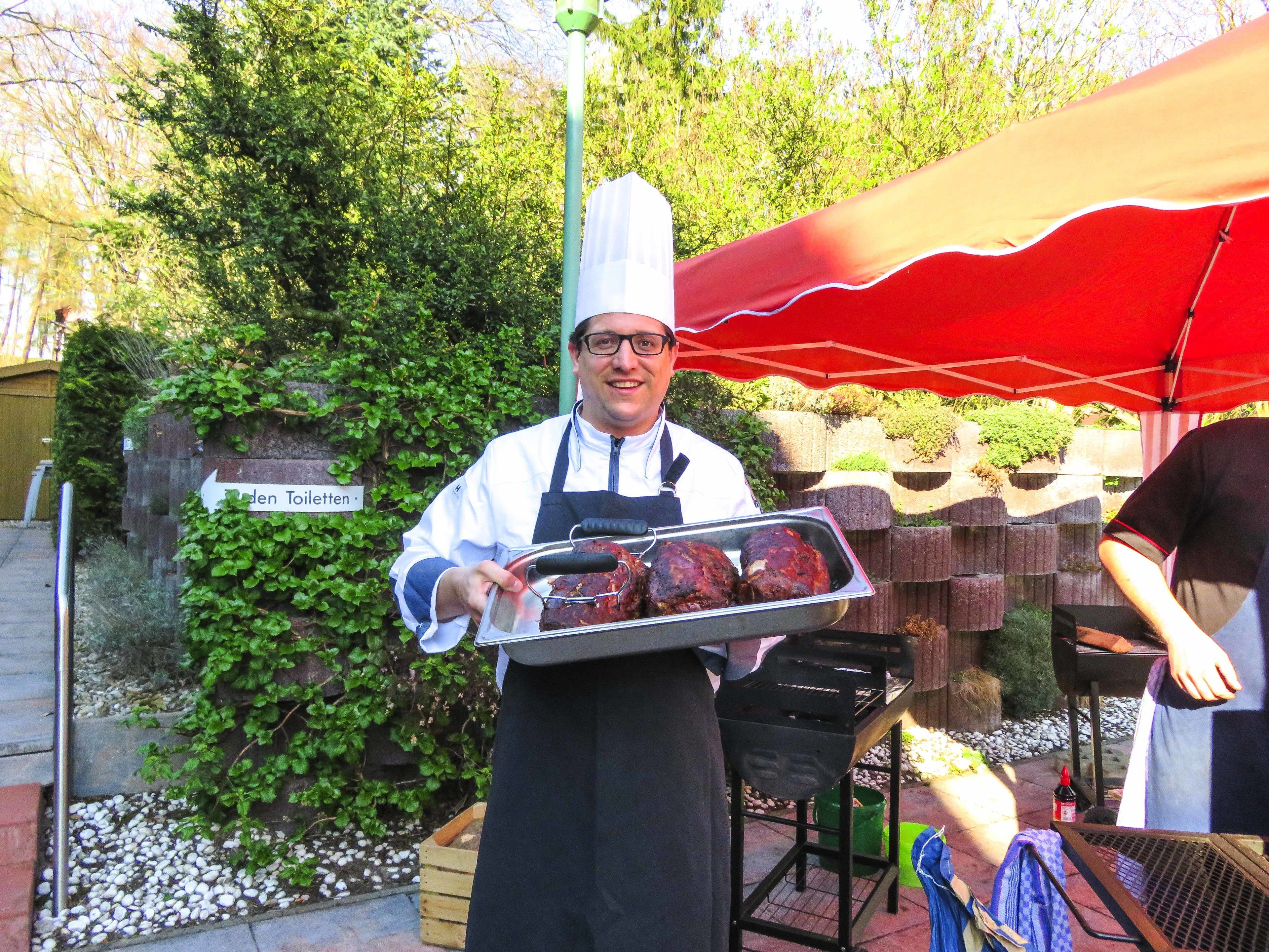 A chef holding a tray of meat under a red umbrella, with a sign reading "Garden Toiletten" in the background.