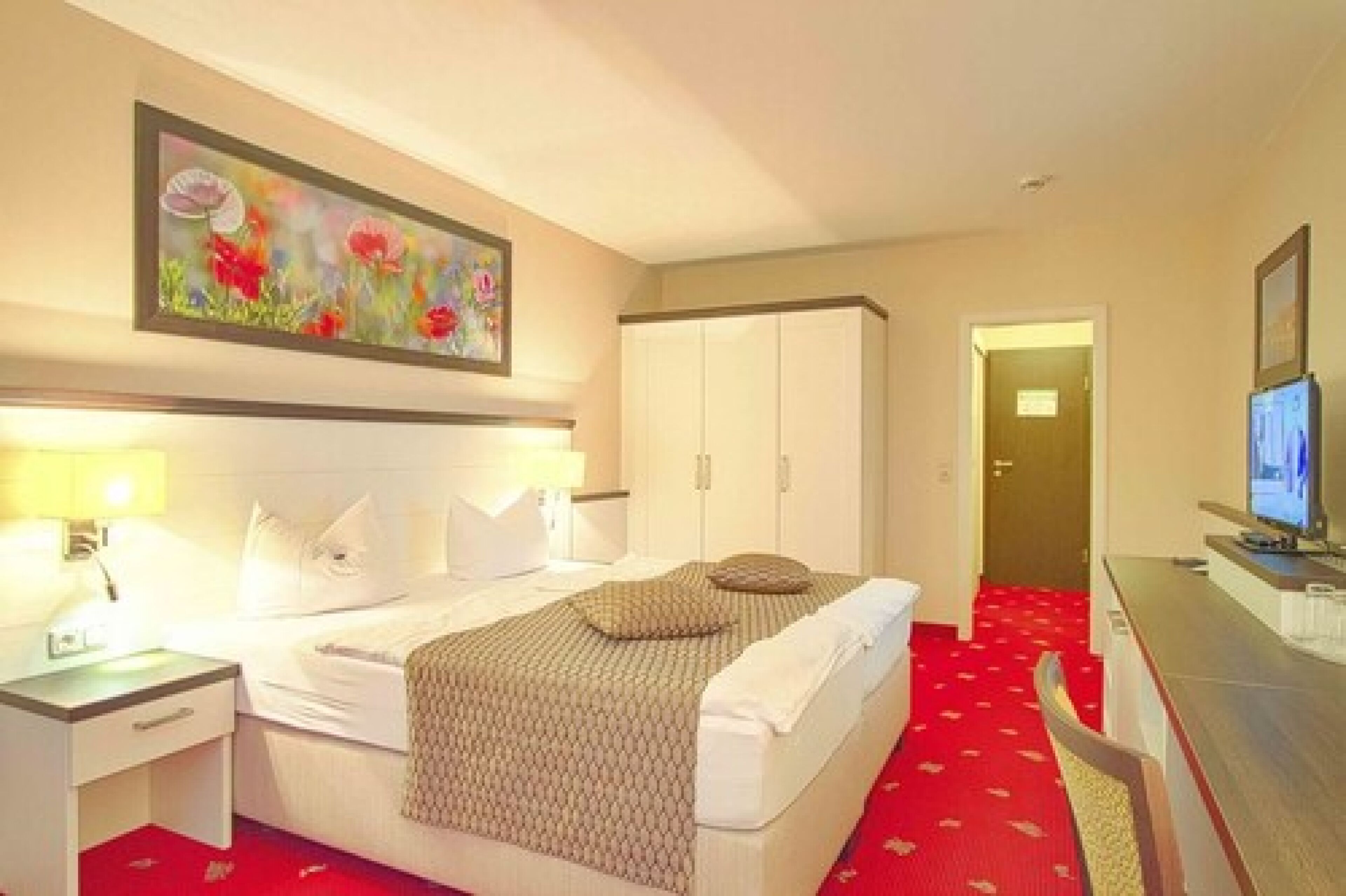 A hotel room with a bed, bedside table, lamp, TV, and a painting on the wall.