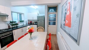 A modern kitchen with white cabinets, a black countertop, and a stainless steel refrigerator. There is a dining table with a white tablecloth and red chairs. A vase with flowers is on the table. - TopHome Finsbury Park (London)
