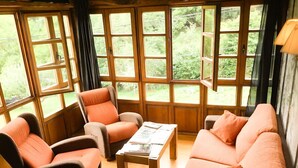 Una habitación acogedora con piso de madera, un sofá, dos sillones y una mesa de centro. Ventanas amplias con cortinas ofrecen vistas al exterior, lleno de vegetación.