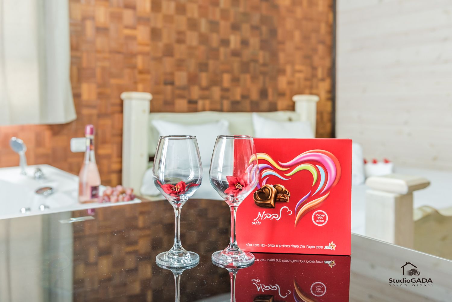 A hotel room with a wooden wall, a bed with white bedding, and a table with two wine glasses and a red box.
