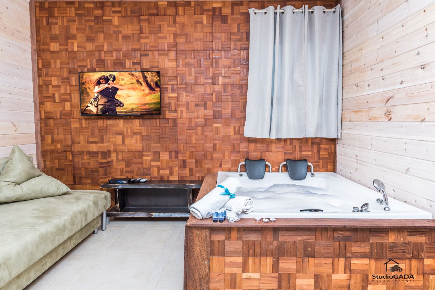 Quarto com parede de madeira em padrão herringbone, cama com duas almofadas, televisão fixada na parede, um banco de madeira e uma pequena mesa com uma toalha e uma chávena.