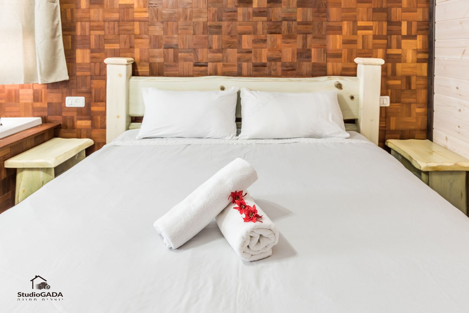 A hotel room with a wooden herringbone-patterned headboard, a neatly made bed with white linens, and a rolled towel with red flowers on the bed.