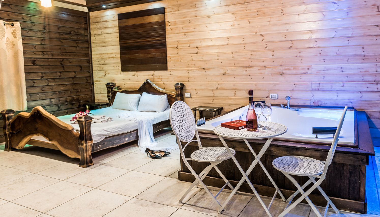 Una habitación de cabaña de madera con una cama, una mesita con una botella y vasos, y un jacuzzi.