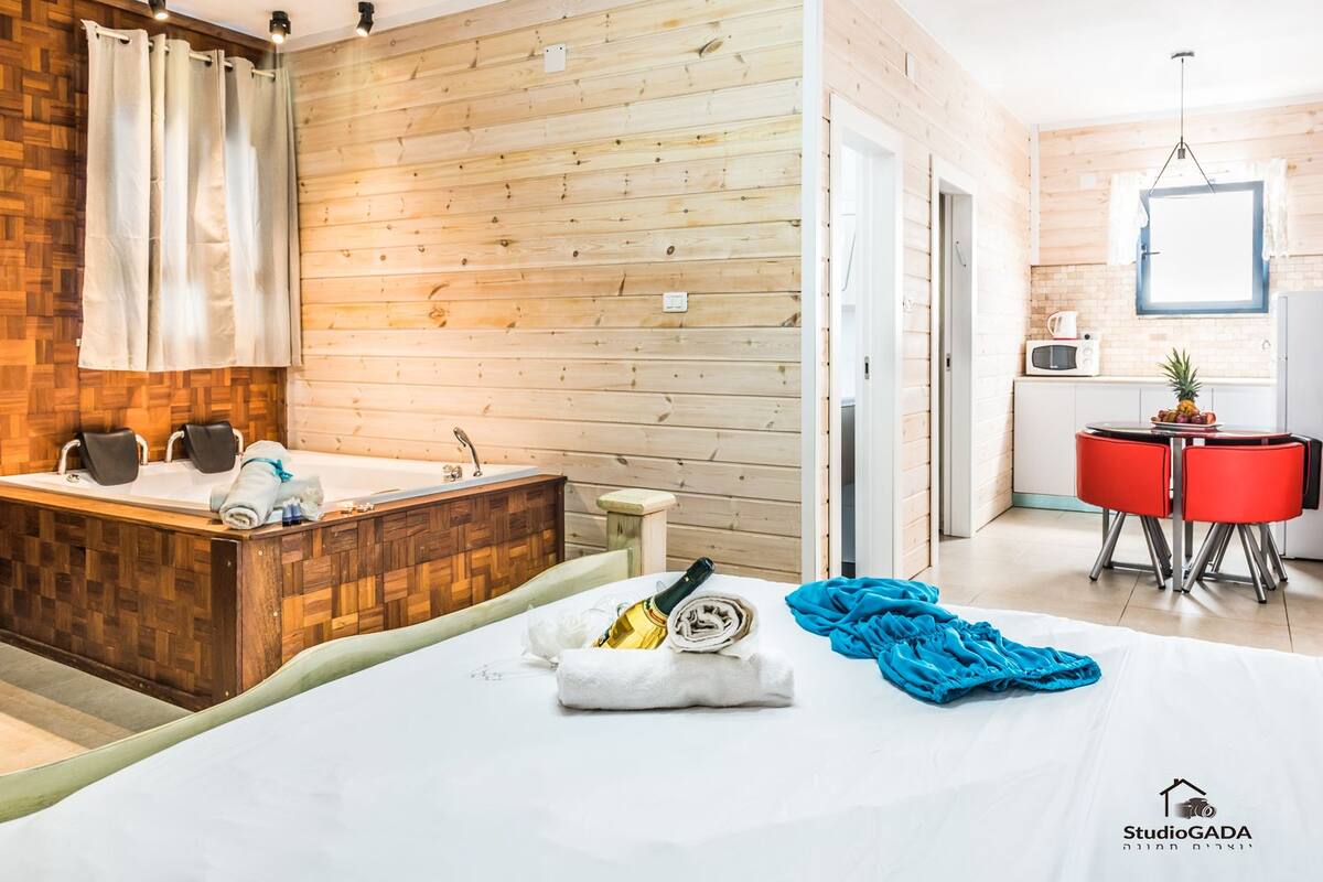 A hotel room with a wooden wall, a bathtub, a bed with towels, a small table with a fruit bowl, and a red chair.