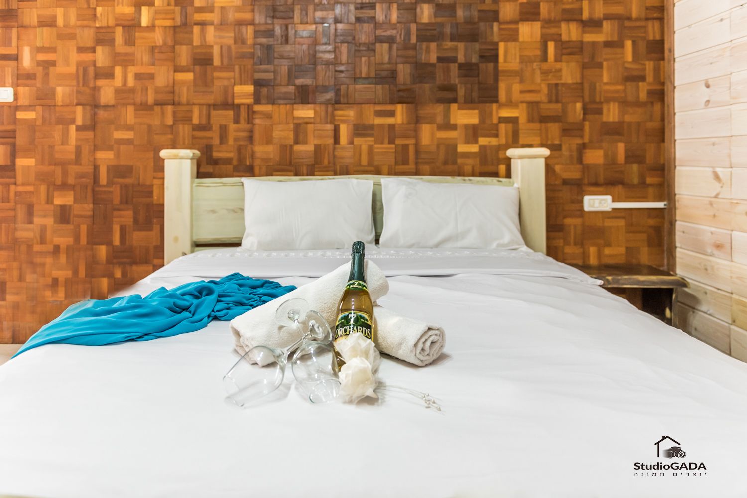 Un lit bien fait, avec du linge de lit blanc, une serviette bleue et une bouteille de vin Orchard's. La chambre est dotée d'un mur en bois avec un motif en chevrons et d'une tête de lit blanche.
