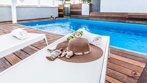 Uma área na beira da piscina com uma espreguiçadeira branca, chapéu de sol, protetor solar e uma garrafa de água.