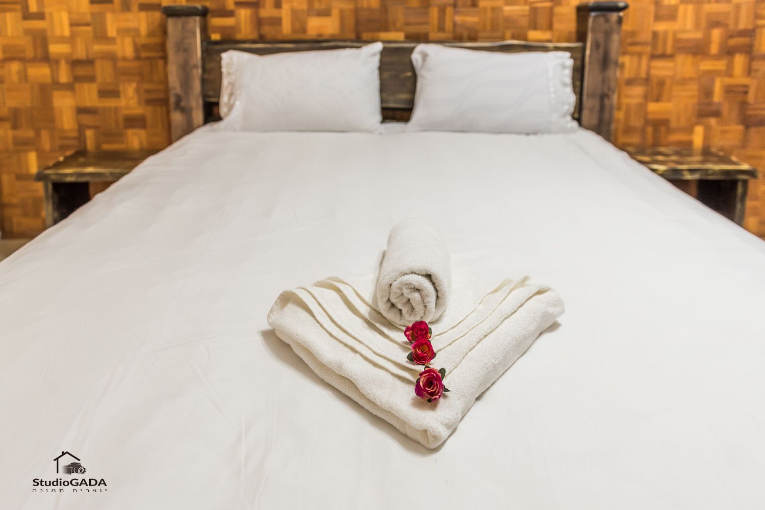 A neatly made bed with a rolled white towel and three red roses on it, set against a wooden headboard and wall.