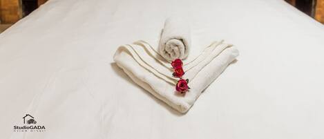 A neatly made bed with a rolled white towel and three red roses on it, set against a wooden headboard and wall.