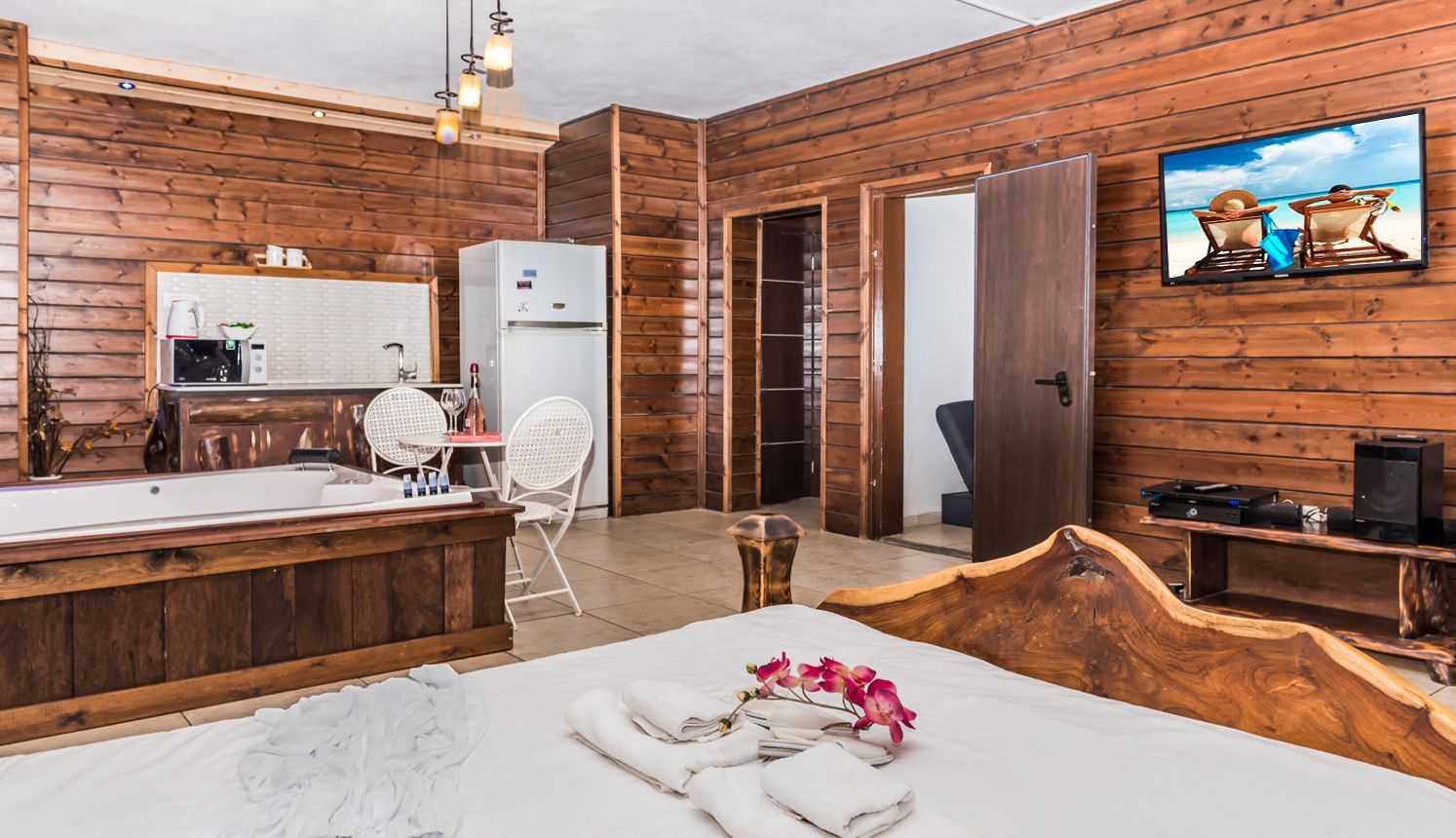 A wooden cabin-style room with a bed, a small kitchenette, a TV, and a door leading to another room.