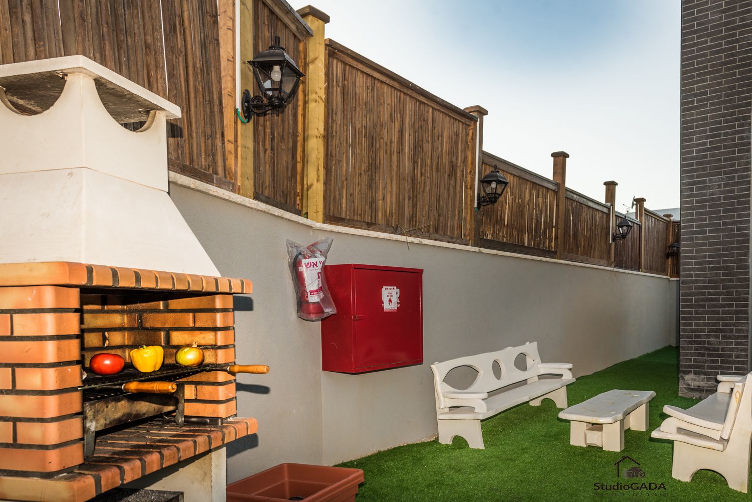 Un barbecue extérieur en briques, agrémenté de tomates et de poivrons, une extincteur rouge et des meubles d’extérieur blancs, le tout posé sur une pelouse artificielle.