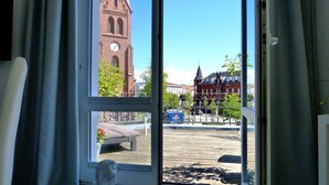 Blick aus einem Fenster mit einer Statue auf dem Sims, einem Glockenturm und einem Park mit Bänken und einem Brunnen.
