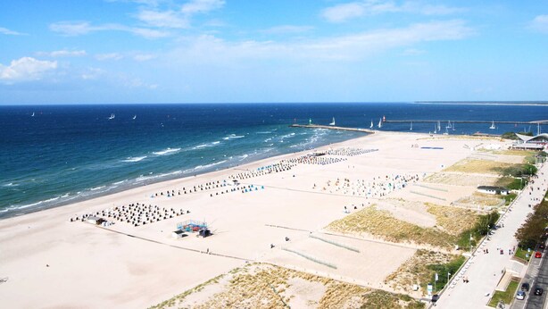 Ein Strand mit einer langen Stegkonstruktion, die ins Meer ragt, ein Sandbereich mit Menschen und ein klarer Himmel.