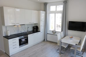 Una cocina moderna con armarios blancos, encimera negra y horno empotrado. Hay una zona de comedor con mesa y sillas blancas, además de una ventana con cortinas.