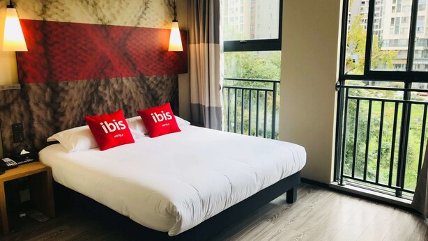 Quarto de hotel com uma cama grande, duas almofadas vermelhas com a inscrição "ibis Hotels", cabeceira com padrão marmorizado, mesa de cabeceira com telefone e varanda com vista para edifícios e vegetação.