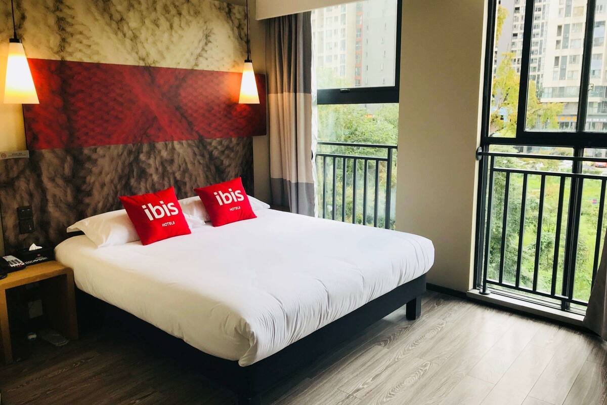 Une chambre d’hôtel avec un grand lit, deux oreillers rouges ornés du texte « ibis Hotels », une tête de lit à motifs de marbre, une table de chevet avec un téléphone et un balcon offrant une vue sur les bâtiments et la verdure.